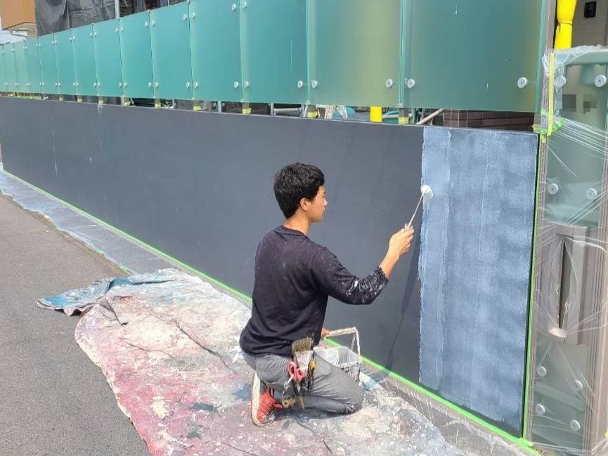 弥富市の外壁塗装におけるコンクリート擁壁（塀）の下塗り作業風景。本塗りの密着性を高めるため、ローラーで下地調整塗料を塗る職人の写真。