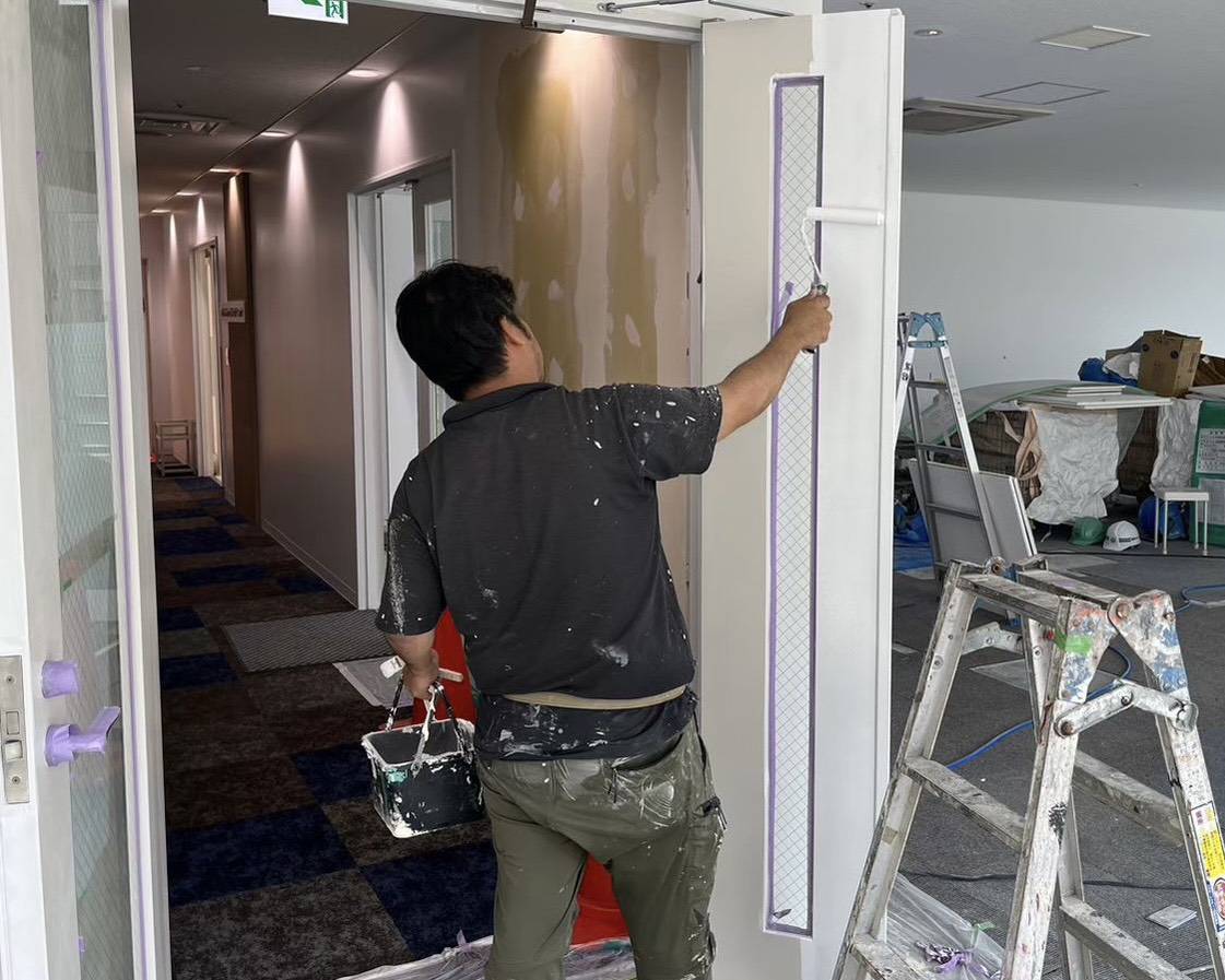 名古屋市のテナントビルオフィス内装塗装におけるドア（扉）のローラー塗り作業風景。ガラス面を綺麗に養生して真っ白に塗り替える職人の写真。