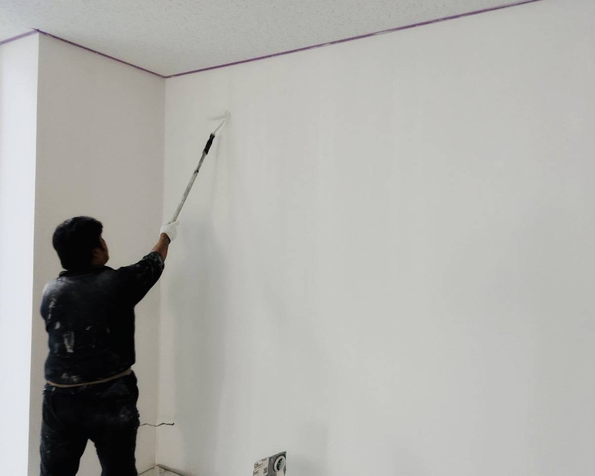 名古屋市のオフィス内装塗装における壁面のローラー塗り作業風景。天井の境目を養生して真っ白な塗料を塗り重ねる職人の写真。