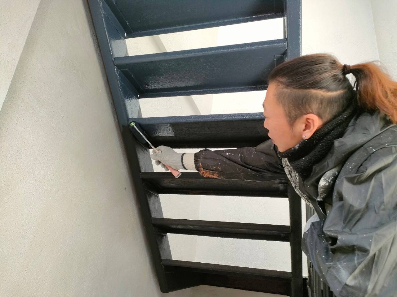 岐阜市のマンション修繕における鉄骨階段裏面の塗装風景。ローラーを使ってクリーンマイルド（75-30D）を塗り重ねる職人の写真。