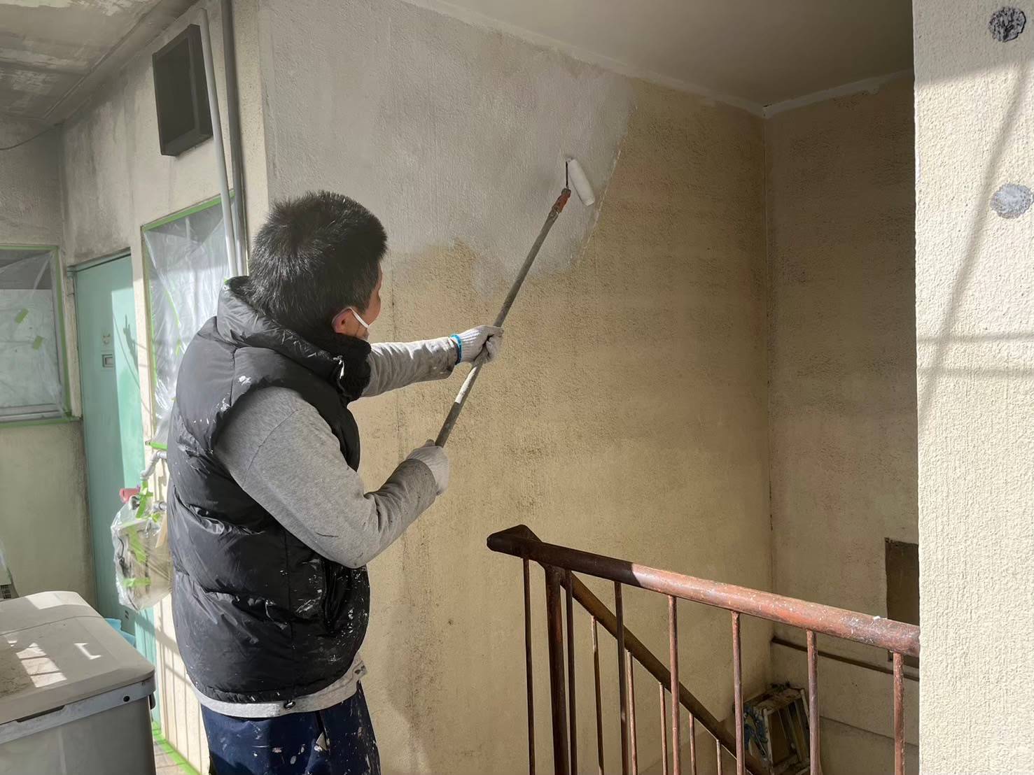 岐阜市のマンション修繕における階段室の壁面塗装風景。長柄のローラーを使って水性セラミシリコン（22-85A）を塗る職人の写真。