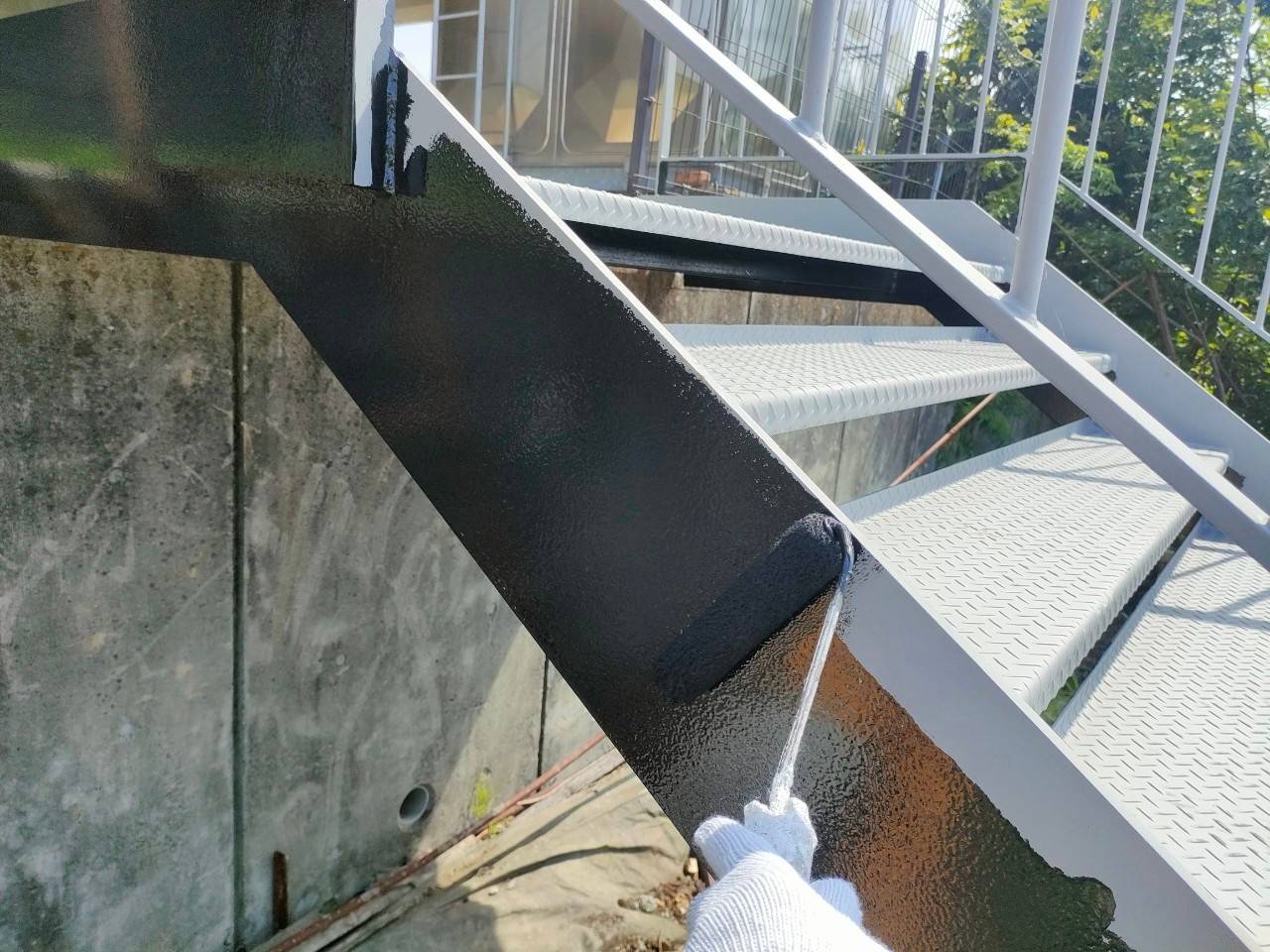 半田市の鉄骨階段塗装における側面ササラ部分のローラー塗り作業風景。グレーの下地から黒色へ塗り替える職人の手元写真。